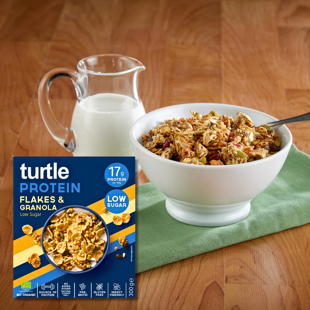 Bowl of Turtle Protein Flakes & Granola with a jug of milk and the cereal box on a wooden table.